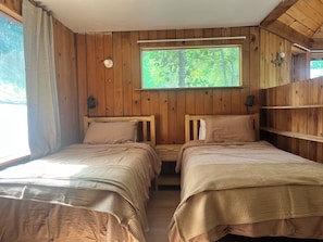 Una cabana amb dos llits de fusta, una finestra amb vista als arbres i una estanteria amb una làmpada.
