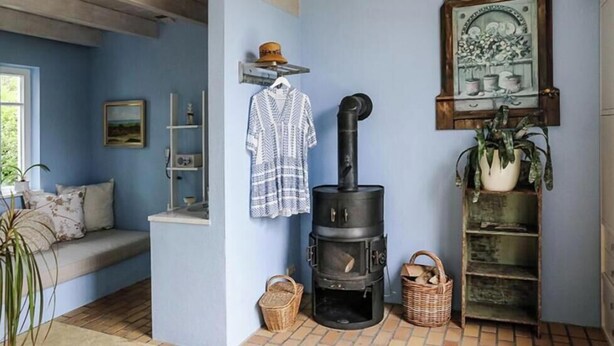 Una habitación acogedora con pared azul, un banco de madera, un estufa de leña negra, un estante con planta en maceta y un cuadro enmarcado.