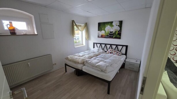 Ein Schlafzimmer mit einem Doppelbett, einem Heizkörper, einem Fenster mit Vorhängen und einem Wandbild.