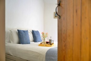 Ein Bett mit weißer Bettwäsche und zwei blauen Kissen, ein Nachttisch mit Kaffeemaschine, einer Tasse und einer kleinen Vase mit Blumen.