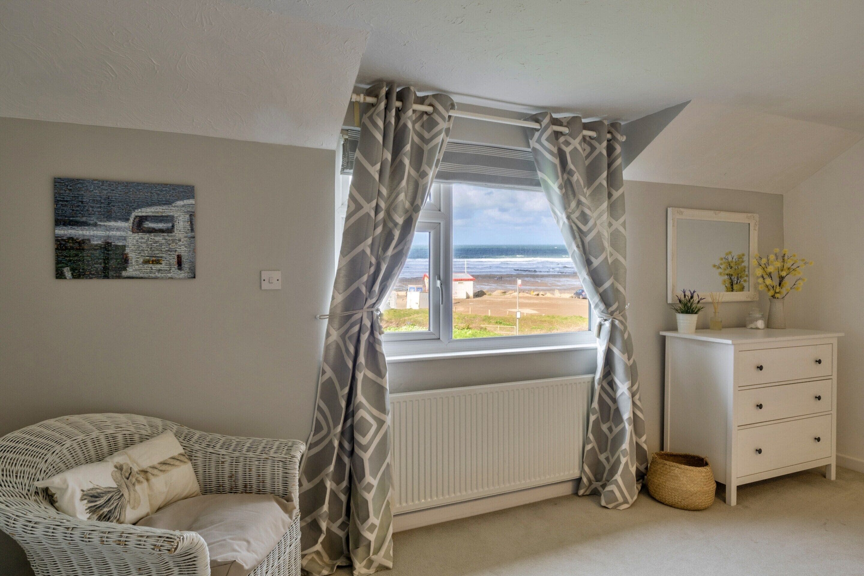 A bedroom with a view of the sea, a white dresser, a wicker chair, and a radiator.