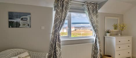 A bedroom with a view of the sea, a white dresser, a wicker chair, and a radiator.