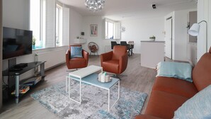 A modern living room with a television, sofa, armchair, and a dining area in the background. - Sun-facing Apartment With Ijsselmeer Views (Makkum)