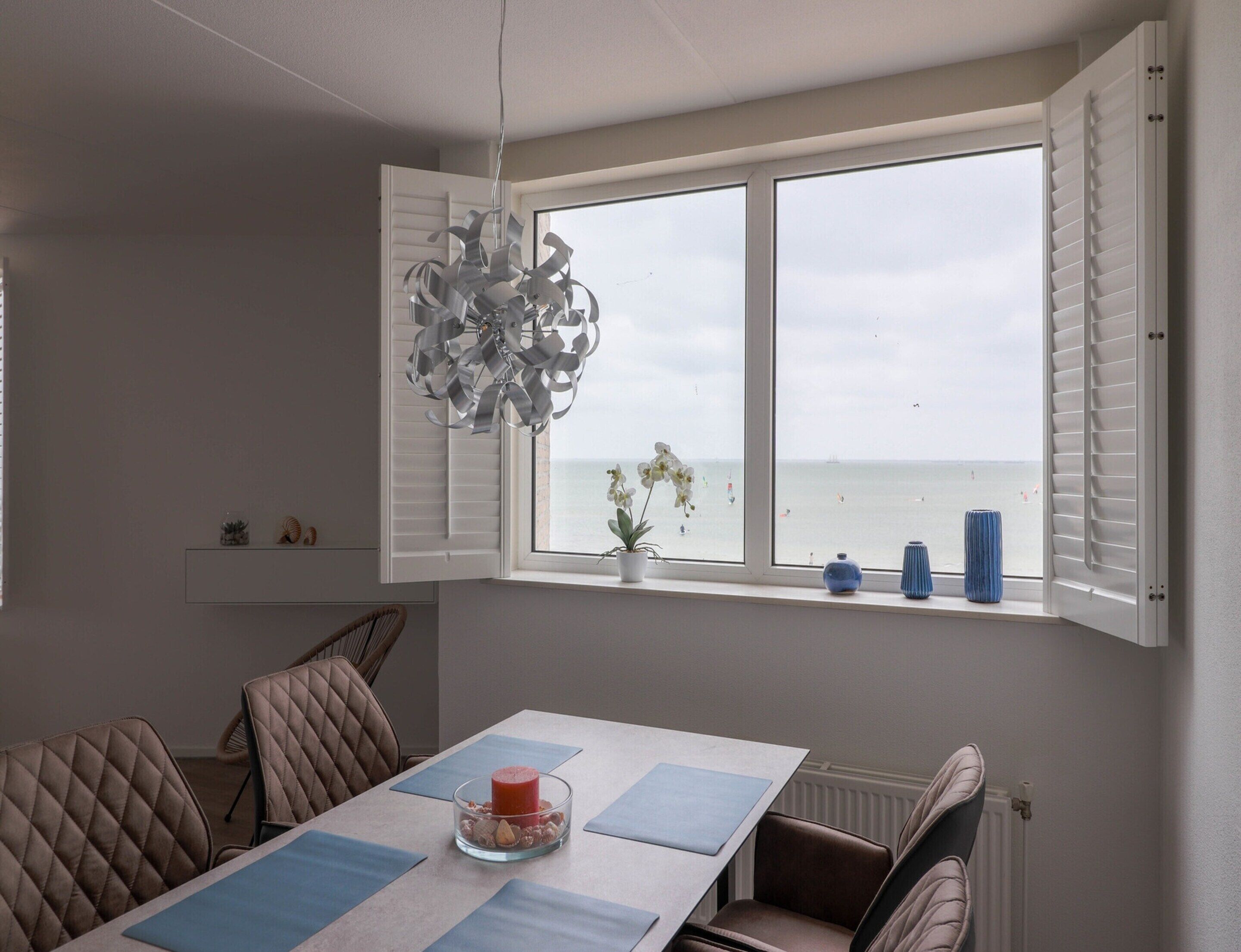 Un comedor con una mesa, sillas, una lámpara colgante, vista al mar a través de una ventana y un estante con adornos.