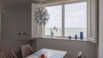 Un comedor con una mesa, sillas, una lámpara colgante, vista al mar a través de una ventana y un estante con adornos.