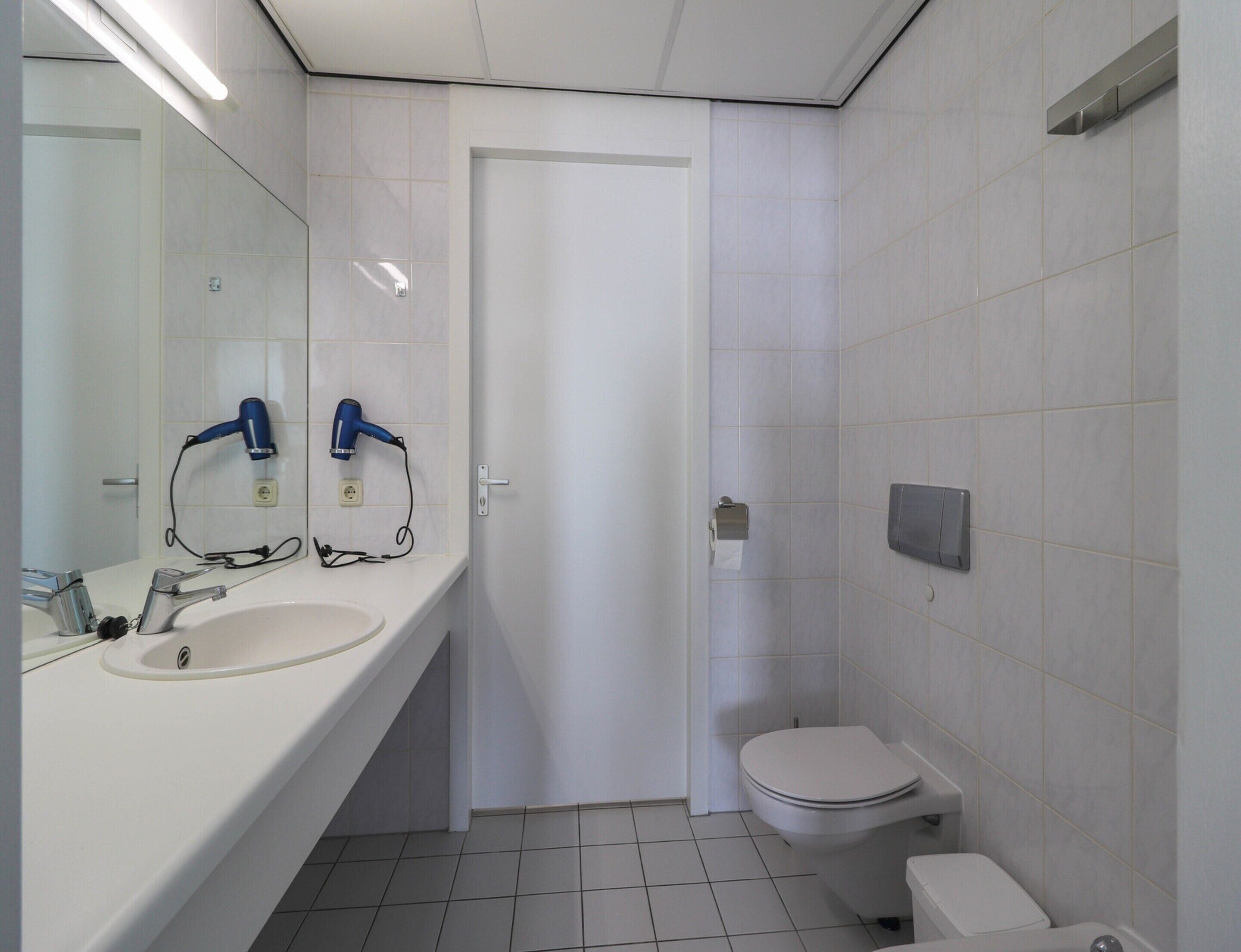 Een badkamer met een witte wasbak, twee haardrogen, een toilet en een spiegel.