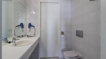 Een badkamer met een witte wasbak, twee haardrogen, een toilet en een spiegel.
