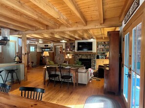 A cozy cabin interior with a stone fireplace, wooden ceiling beams, a television mounted above it, and a bookshelf filled with books.