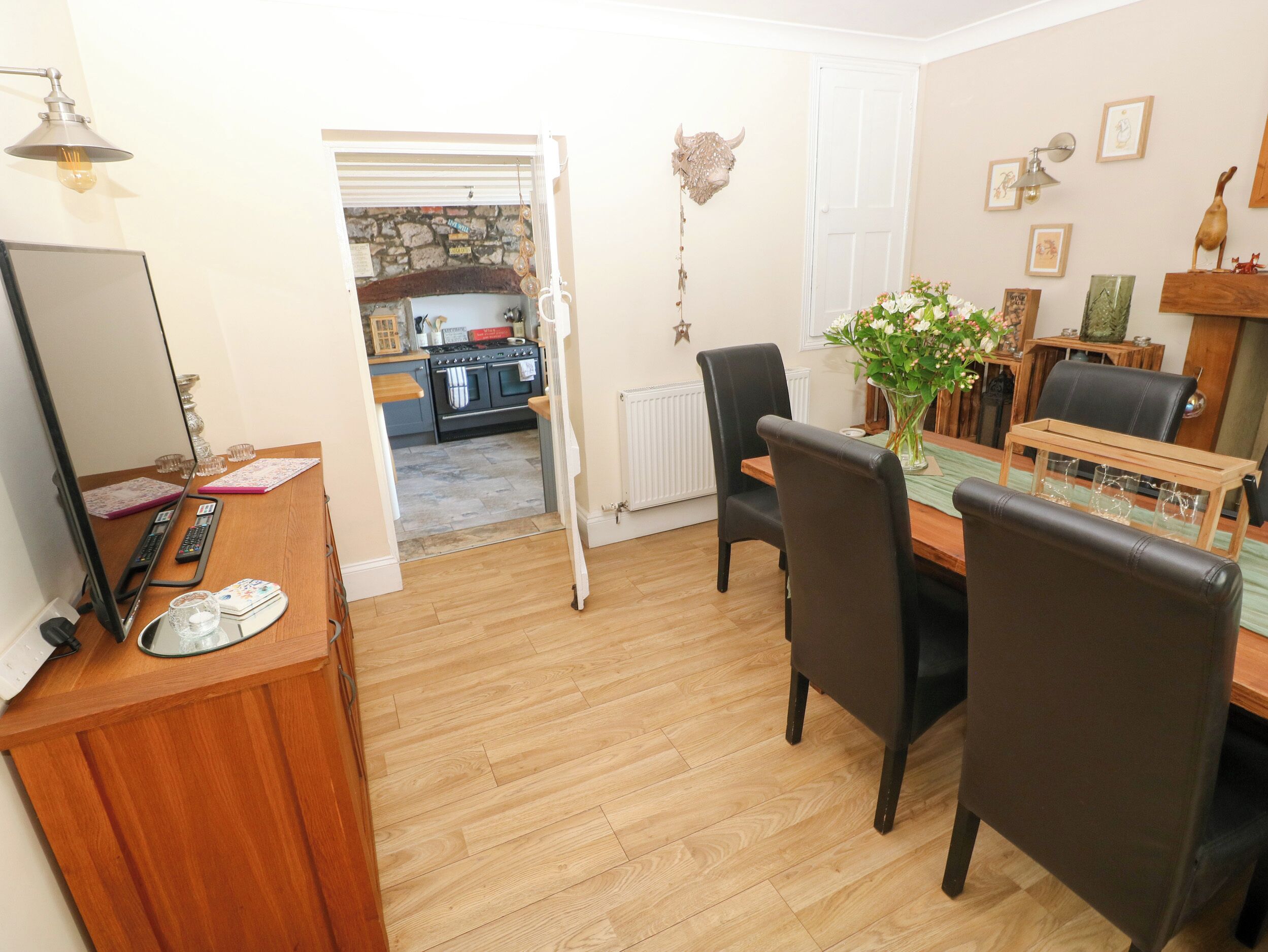 A dining area with a wooden table, chairs, and a vase with flowers. A kitchen is visible in the background.