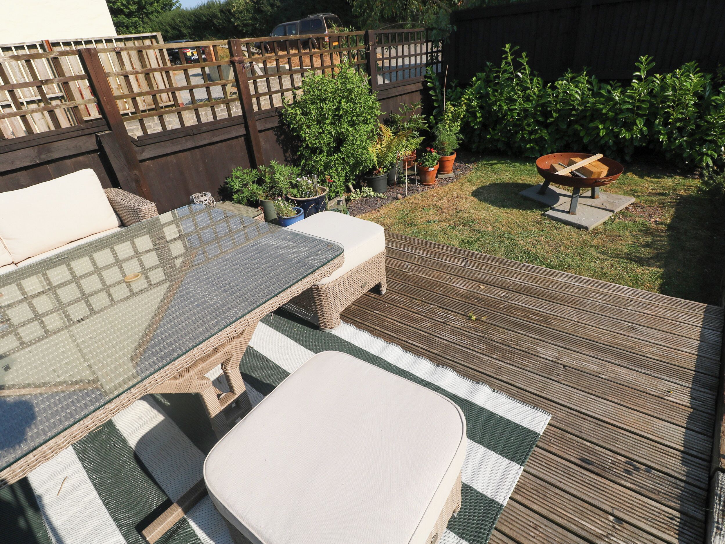 A wooden deck with a glass-top table and cushioned seating, surrounded by potted plants and a wooden fence.