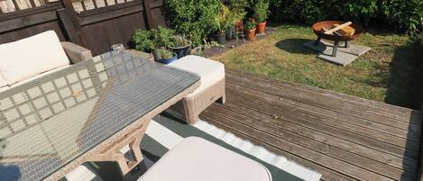 Eine Holzdielen-Terrasse mit Tischplatte aus Glas und gepolsterten Sitzgelegenheiten, umgeben von Blumentöpfen und einem Holzzaun.