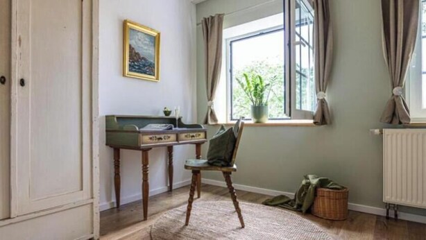 Una habitación con escritorio, silla, ventana con cortinas, radiador y una planta pequeña.