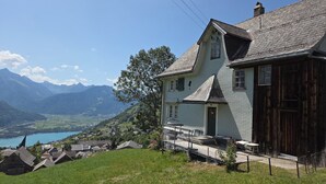 Ein zweistöckiges Haus mit Balkon, umgeben von üppiger, grüner Landschaft und Bergen im Hintergrund.