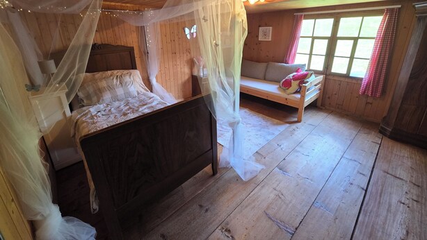 Ein Zimmer aus Holz mit einem Bett, einer Bank und einem Fenster mit Vorhängen.