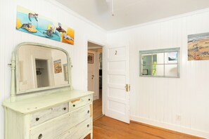Ein Zimmer mit weißer Tür, einem Schrank mit Schubladen und einem Spiegel. An den Wänden befinden sich Gemälde.