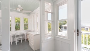 Une pièce avec un bureau blanc, une chaise et des étagères intégrées. De nombreuses fenêtres laissent entrer la lumière naturelle et offrent une vue sur l’extérieur.