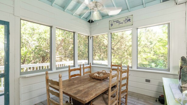 Une véranda couverte avec une table et des chaises en bois, de grandes fenêtres et un ventilateur de plafond.
