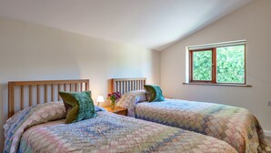 A room with two beds, each with a wooden headboard, a nightstand with a lamp, and a window with a view of greenery. - Stunning Bamburgh Coast Apartment (Okehampton)