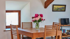 A dining area with a wooden table, chairs, a vase of flowers, a painting on the wall, and a bookshelf with books and a bowl. - Stunning Bamburgh Coast Apartment (Okehampton)