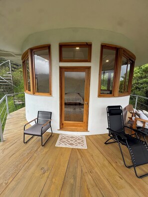 A covered outdoor area with a wooden deck, two chairs, a small table, and a door leading to a bedroom.