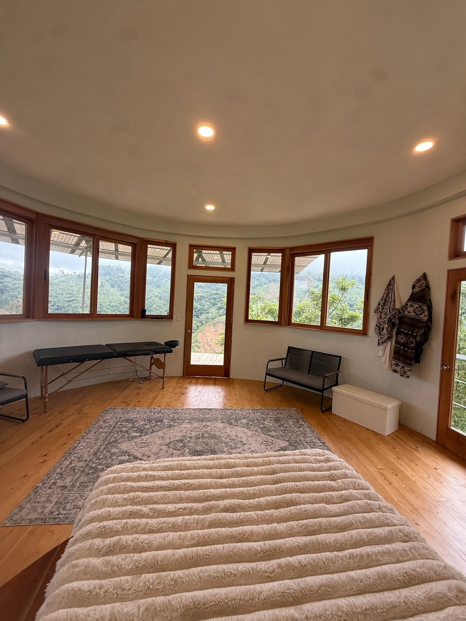 A room with a large bed, a bench, a massage table, and a view of a mountainous landscape through large windows.