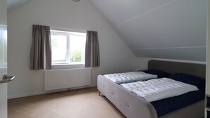 A room with a sloped ceiling, a bed with white bedding and blue pillows, a window with curtains, and a radiator. - Private Bungalow With Jetty for 6 (Makkum)