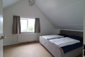 Ein Zimmer mit schräge Decke, einem Bett mit weißer Bettwäsche und blauen Kissen, einem Fenster mit Vorhängen und einem Heizkörper.