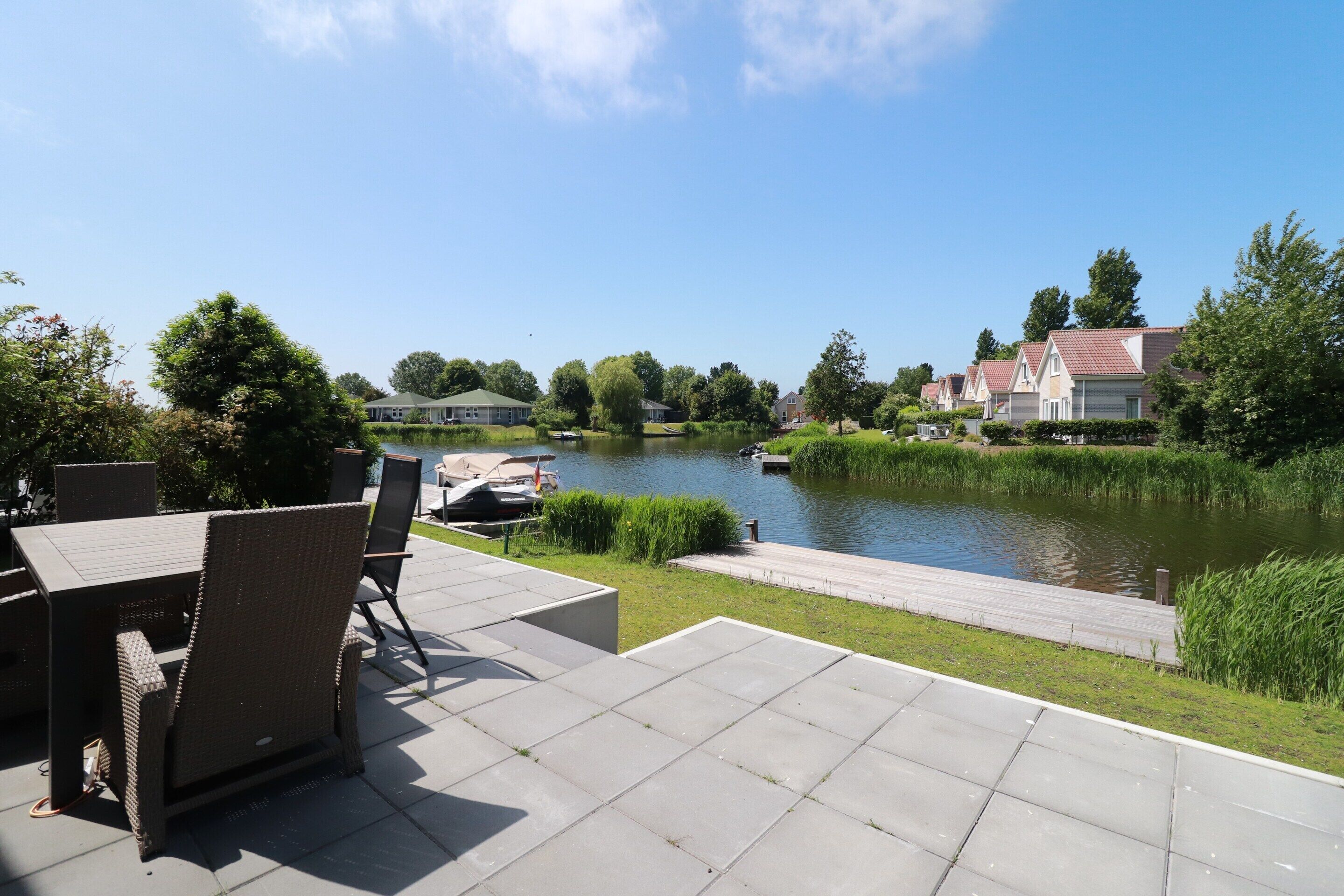 A riverside patio with outdoor furniture, a wooden dock, and houses in the background.