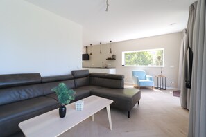A modern living room with a sectional sofa, a coffee table, a blue armchair, and a view of greenery through a window. - Private Bungalow With Jetty for 6 (Makkum)