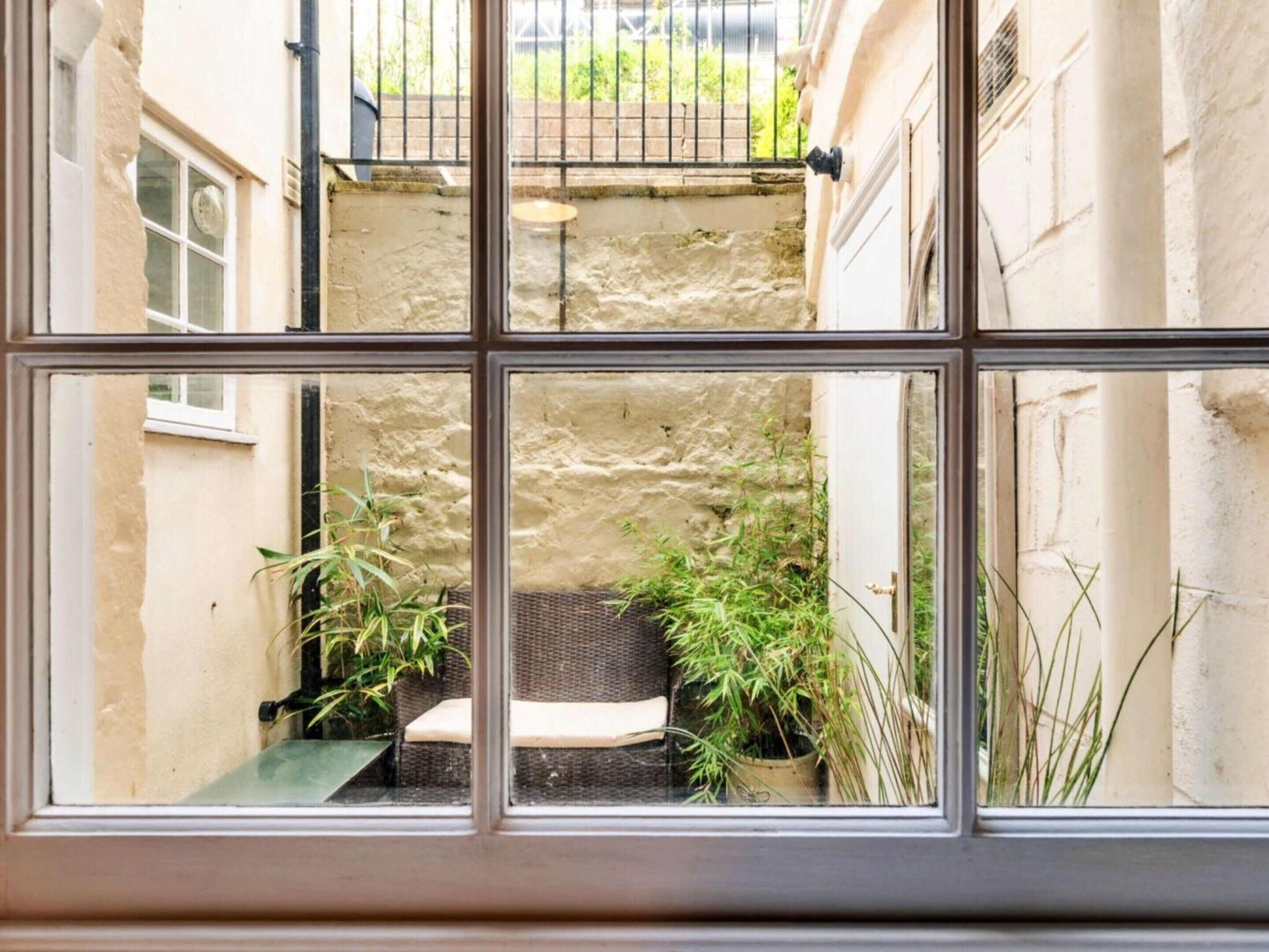 A view through a window of a courtyard with a stone wall, a wicker bench, and potted plants.
