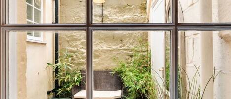A view through a window of a courtyard with a stone wall, a wicker bench, and potted plants.