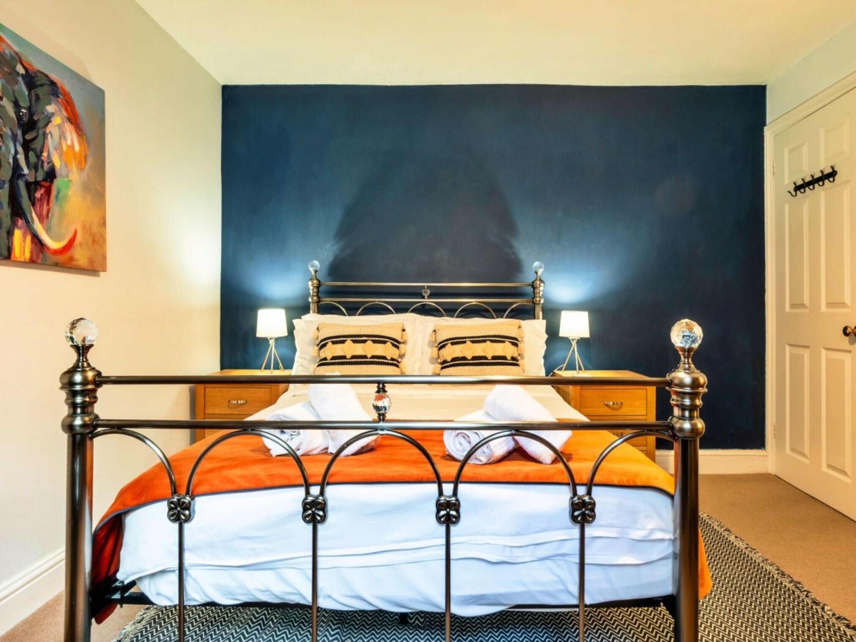 A bed with a metal frame and a dark headboard, a bedside table with a lamp, and a wall with a painting.