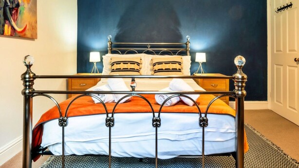 A bed with a metal frame and a dark headboard, a bedside table with a lamp, and a wall with a painting. - Peaceful Dorset Cottage for 4 (Bath)