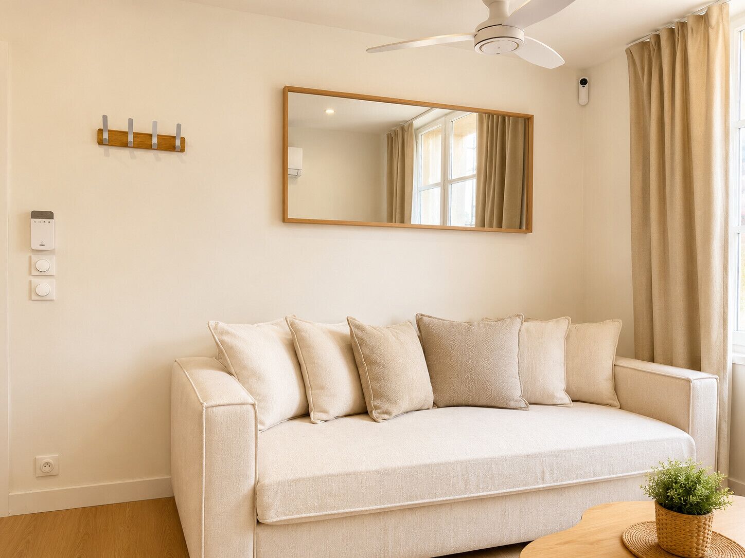 A neutral-colored sofa with multiple cushions, a ceiling fan, a wall-mounted mirror, and a small table with a plant.