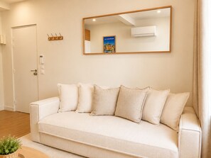 A neutral-colored sofa with multiple cushions, a wall-mounted mirror, a door with a handle, and a small plant on the floor. - Appartement au Panier • Clim • 4 pers (Marseille)