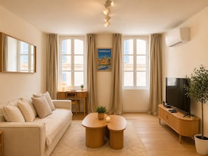 Ein modernes Wohnzimmer mit einer Couch, einem Fernseher, einem Esstisch mit Stühlen und einem großen Fenster mit Vorhängen.
