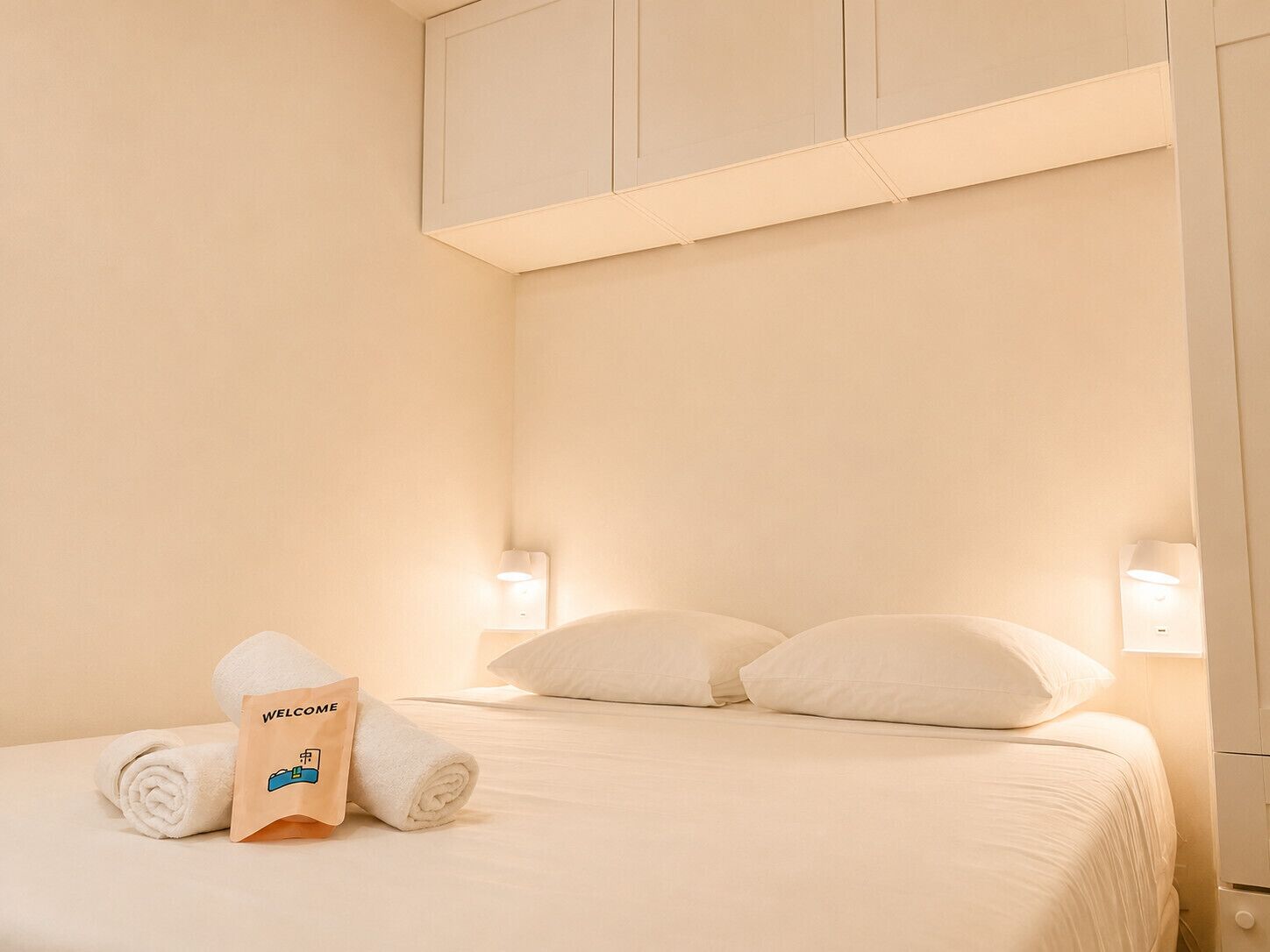 A hotel room with a bed, pillows, a rolled towel, a welcome bag, and wall-mounted lights.