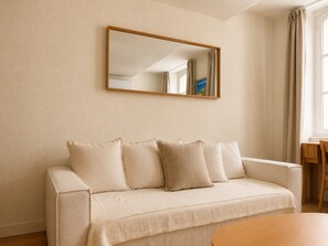 A neutral-colored sofa with cushions, a wooden-framed mirror, and a wooden table in a room with beige walls. - Bail mobilité - Bel appart 1 ch Panier avec clim (Marseille)
