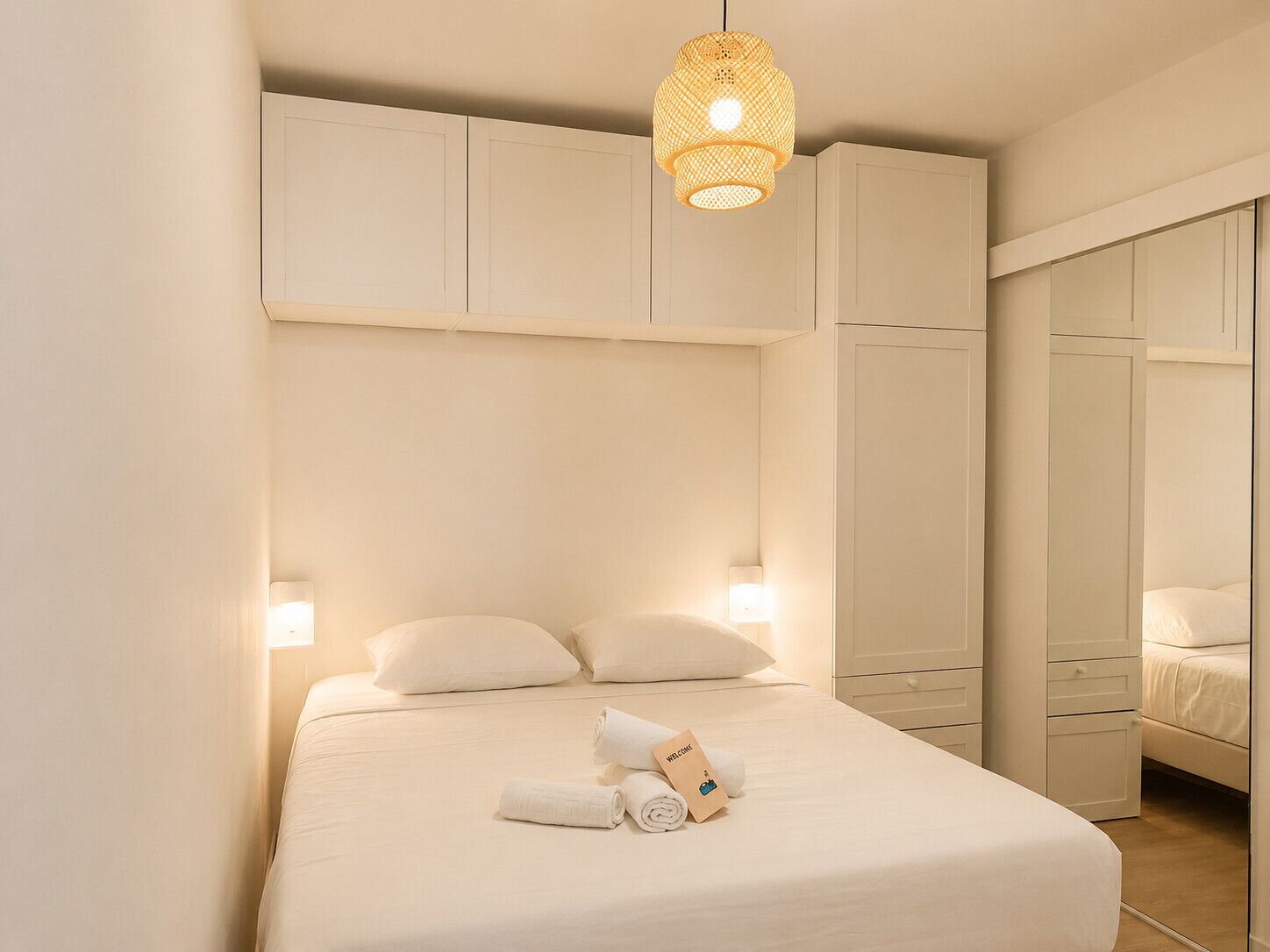 A hotel room with a bed, pillows, a bedside lamp, a hanging light fixture, and a wardrobe.