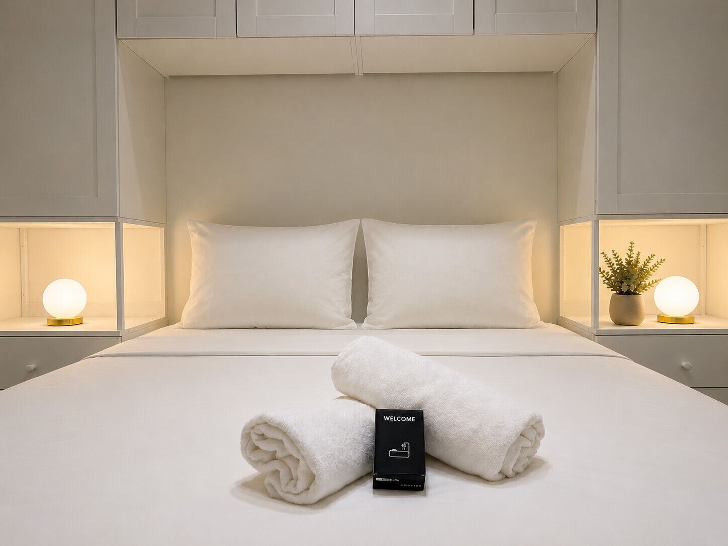 A neatly made bed with white linens and pillows, a rolled towel, a welcome box, and bedside lamps with spherical light fixtures.