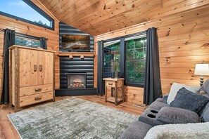 L’intérieur d’un chalet chaleureux, avec une cheminée, des murs en bois, un canapé et une vue sur les arbres par les fenêtres.