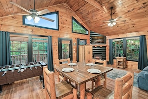 Interior de uma cabana de madeira com uma mesa de jantar, cadeiras, mesa de futebol e lareira.
