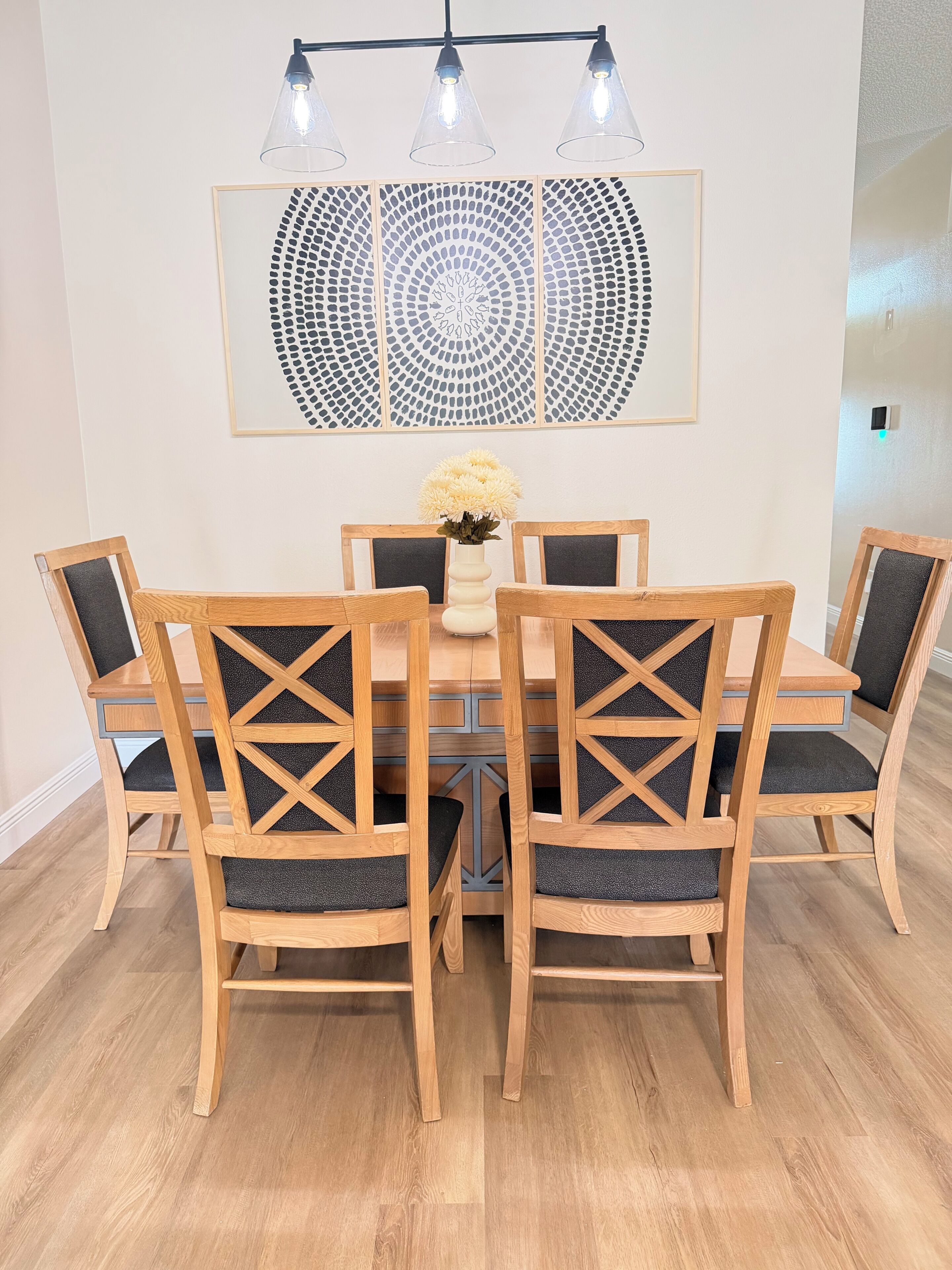 Un comedor con una mesa redonda, seis sillas de madera con respaldo de escalera, y un florero con flores.