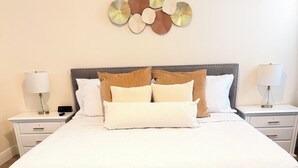 Cama bem arrumada com colcha branca, com duas mesas de cabeceira com abajures, e uma obra de arte na parede acima.