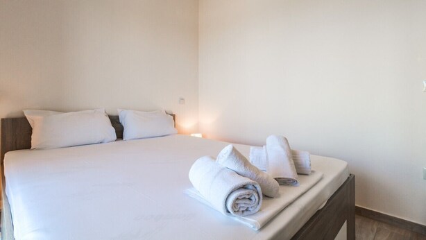 Een netjes opgemaakt bed met witte lakens en kussens, een nachtkastje met opgerolde handdoeken, en een wandlamp.