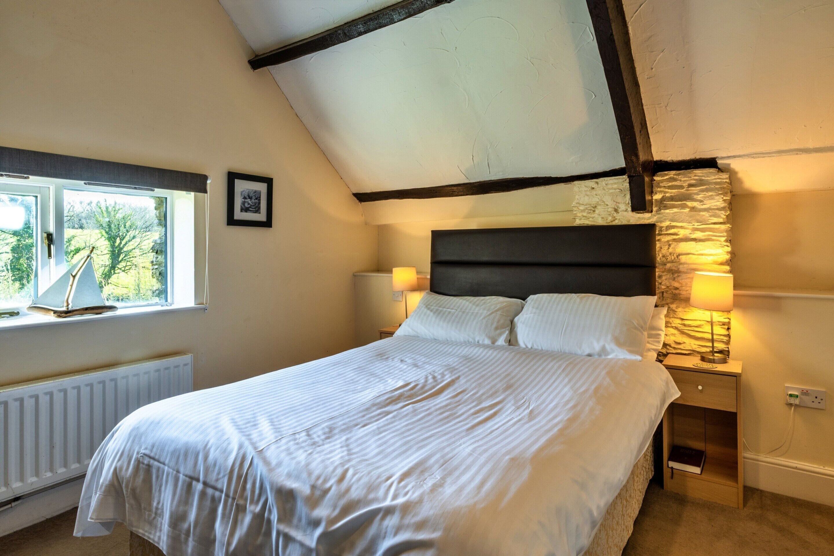 Un dormitorio con cama, ventana, un cuadro en la pared y una mesita de noche con lámpara.