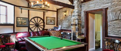A billiards room with a pool table, leather chairs, a stone wall, and a wooden ceiling.