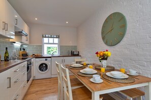 A modern kitchen with a built-in washing machine, a dining area with a wooden table and chairs, and a large wall clock. - Scenic Snowdonia Coastal Cabin (Ashbourne)