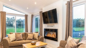 Een moderne woonkamer met een bank, een salontafel, een flatscreen televisie boven een open haard, en grote ramen met uitzicht op een groen landschap.