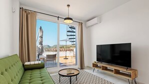 En moderne stue med en grøn sofa, et fladskærms-tv og en stor skydedør, der fører ud til en udendørs terrasse.